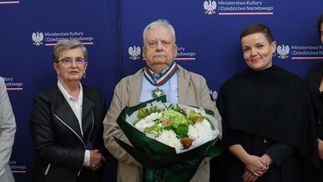 Andrzej Sapkowski wears a medal and holds a bouquet of flowers, flanked by Polish government officials.